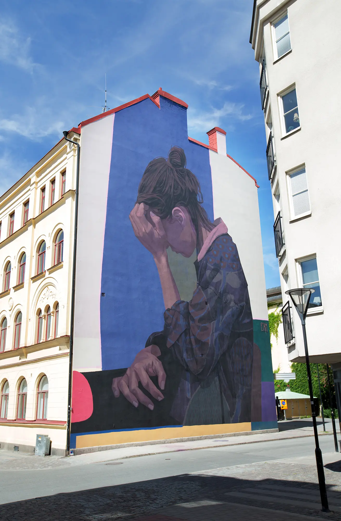På en sidan av ett stort hus syns en stor väggmålning som täcker nästan hela väggen. Målningen föreställer en sittande kvinna som ser nedstämd ut framför en blå bakgrund. Hon lutar sitt huvud mot sin vänstra hand.
