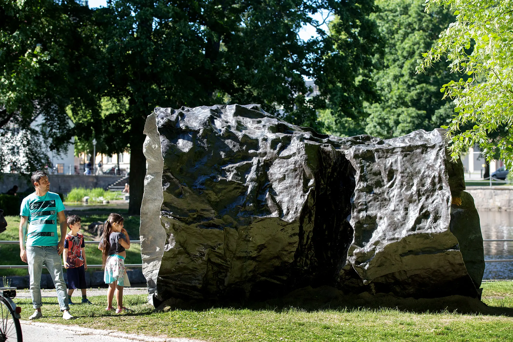 En stor metallskulptur står i en park. Skulpturen har formen av en en ihålig cylinder med ojämn yta. Framför den står en man och två barn och ser på skulpturen.