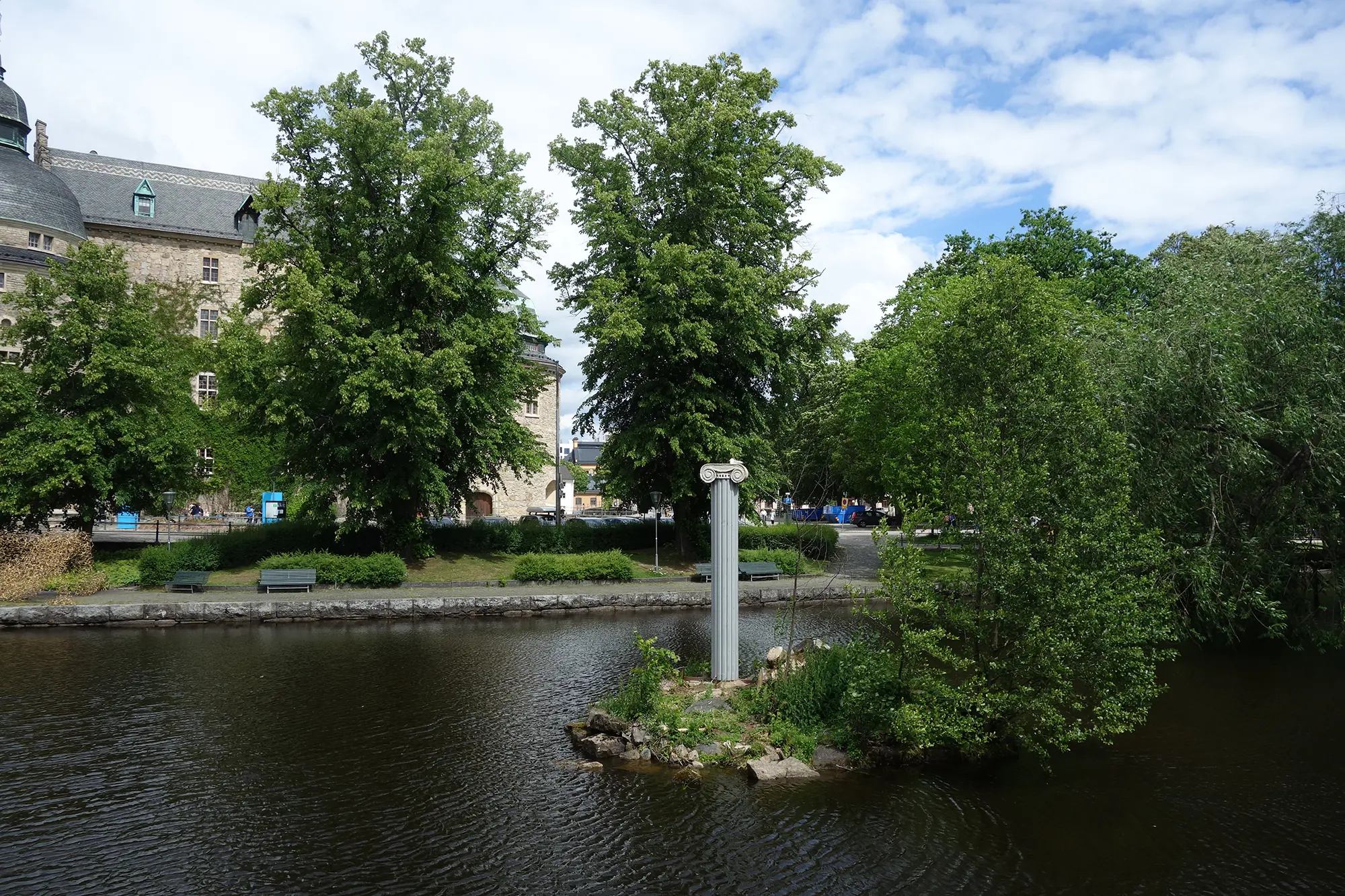Ute i en å syns en liten ö. På ön står en romersk pelare placerad bredvid gröna buskar . I bakgrunden syns Örebro slott och flera träd.