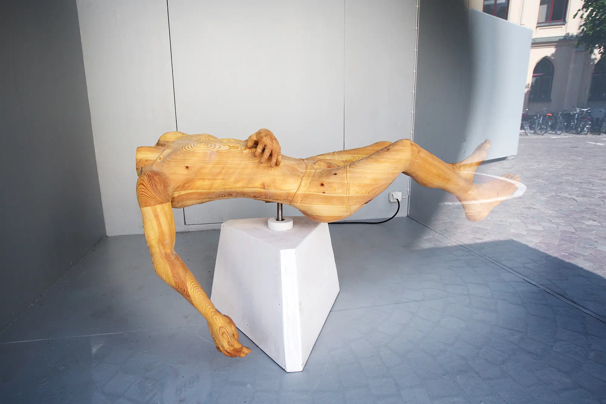 I en glasmonter syns en skulptur av trä. På en ljusgrå träplattform balanserar en liggande människoformad skulptur utan huvud på en plattform. Träet är ljusgult och skulpturen vilar den ena handen på magen och den andra hänger ned mot golvet.
