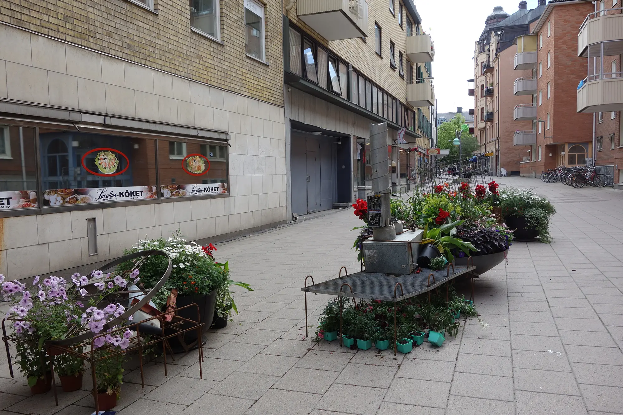 På en lång gata står två stora färgglada blomsterarrangemang placerade. Arrangemangen består av utspridda krukväxter och olika slags metallobjekt placerade i två grupper.