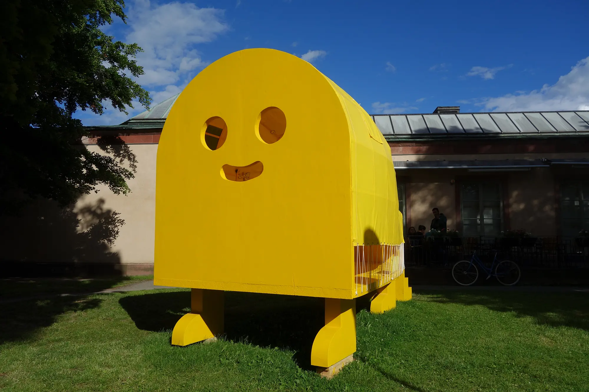 På en gräsmatta framför ett lågt hus står en stor gul skulptur som består av ett avlångt tält med runt tak med två träväggar på kortsidorna. Skulpturen är upphöjd på fyra gula ben. På den ena träväggen är hål utskurna i formen av en glad emoji.