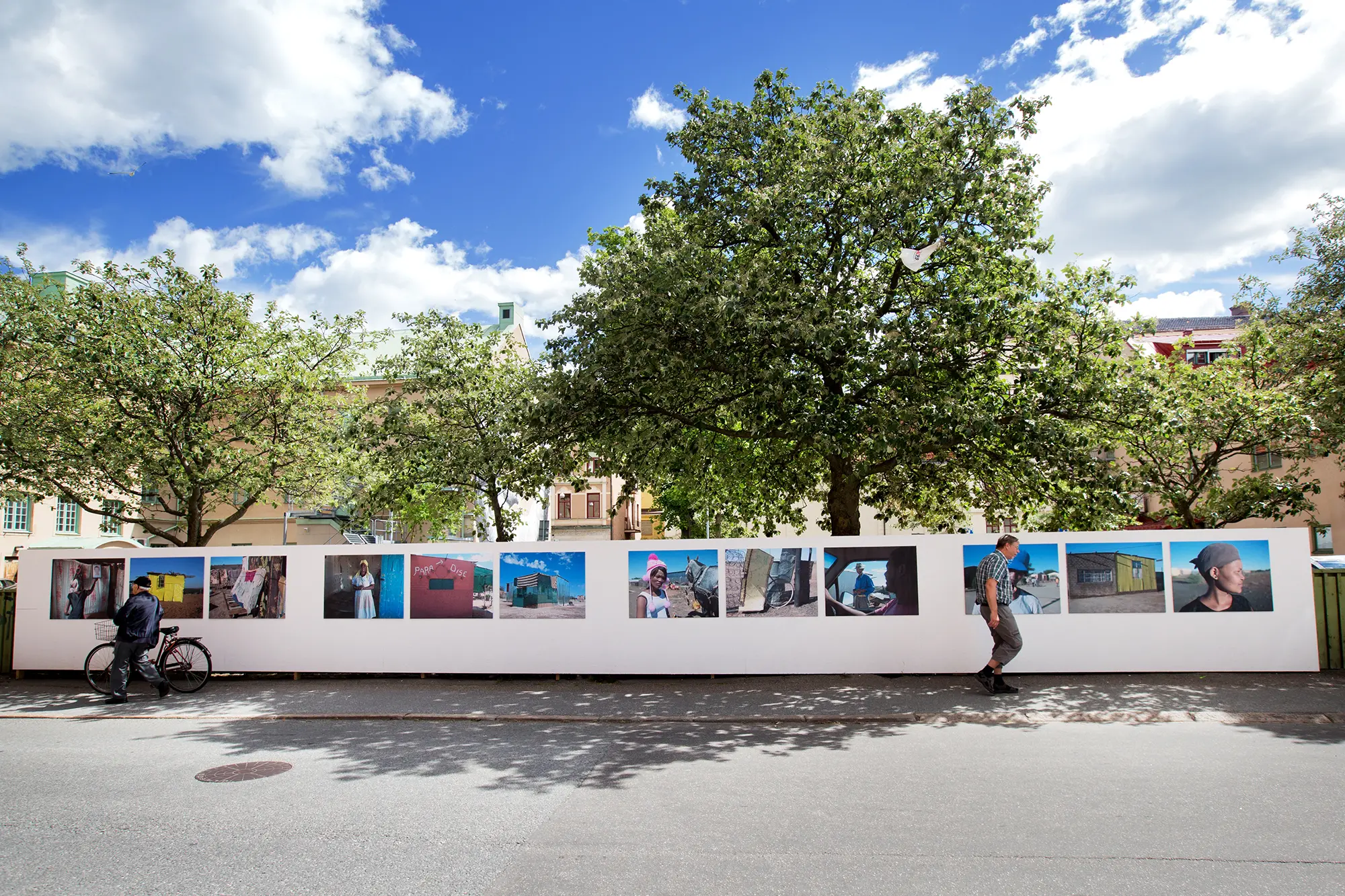 Utomhus på en lång vit vägg framför några träd sitter flera stora färgglada fotografier upphängda på en rad. Bilderna föreställer människor och slitna byggnader i ökenlandskap. På gatan framför går två personer förbi i olika riktningar.