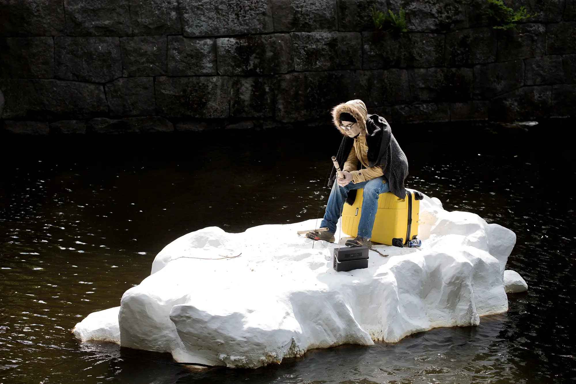 Ute i vattnet i en å är en installation placerad som en ö. Installationen består av en vit snöliknande plattform med en mycket realistisk människoskulptur sittandes på en gul resväska. Figuren bär vinterkläder tittar neråt. I handen håller han ett litet fiskespö.