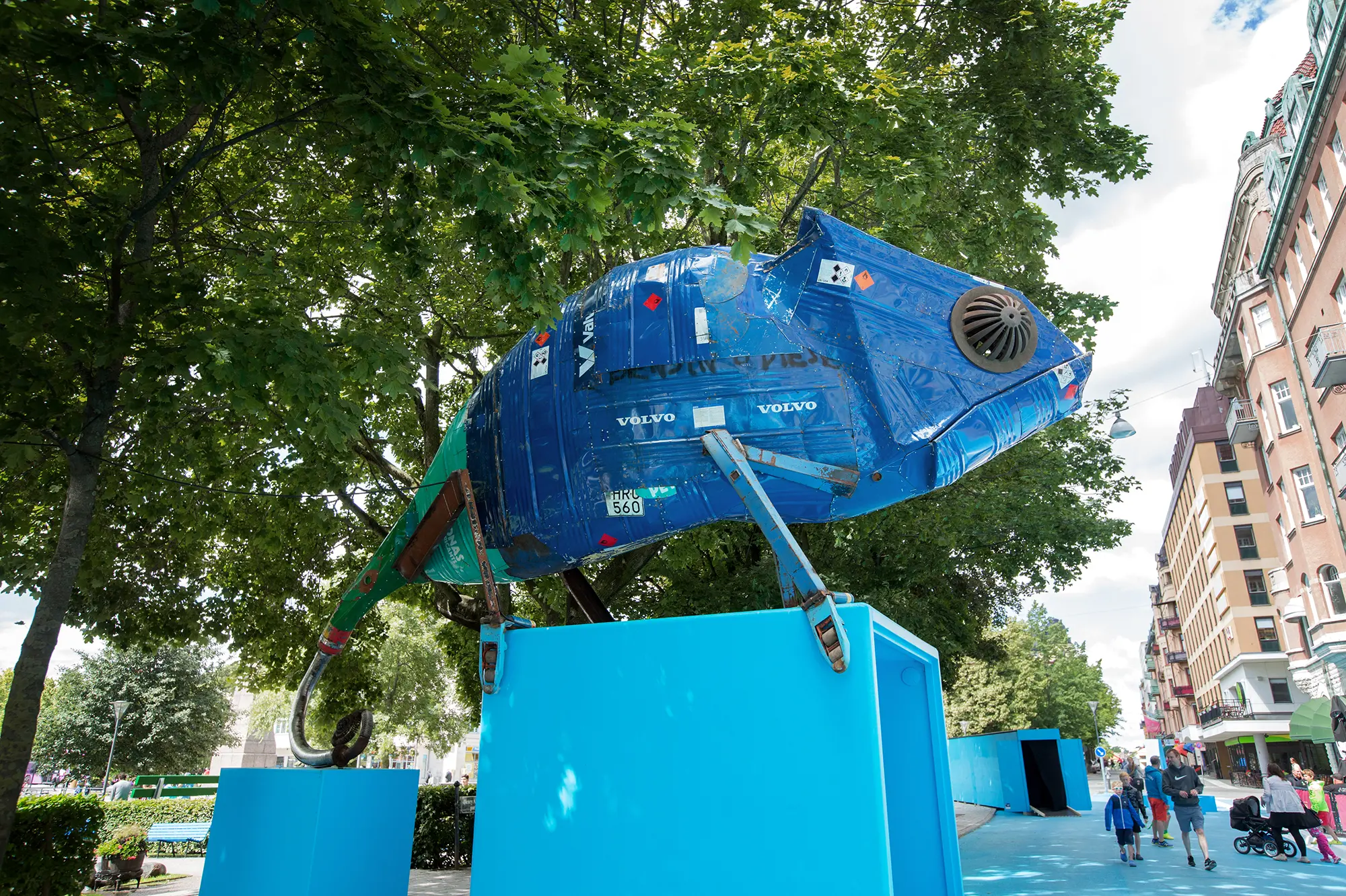 Utomhus uppe på en ljusblå plattform är en stor blå skulptur i metall i formen av en kameleont placerad. Den är uppbyggd av böjda plåtbitar som som sitter nitade som ett lapptäcke. På några av bitarna syns text och symboler, bland annat "VOLVO" och en registreringsskylt
