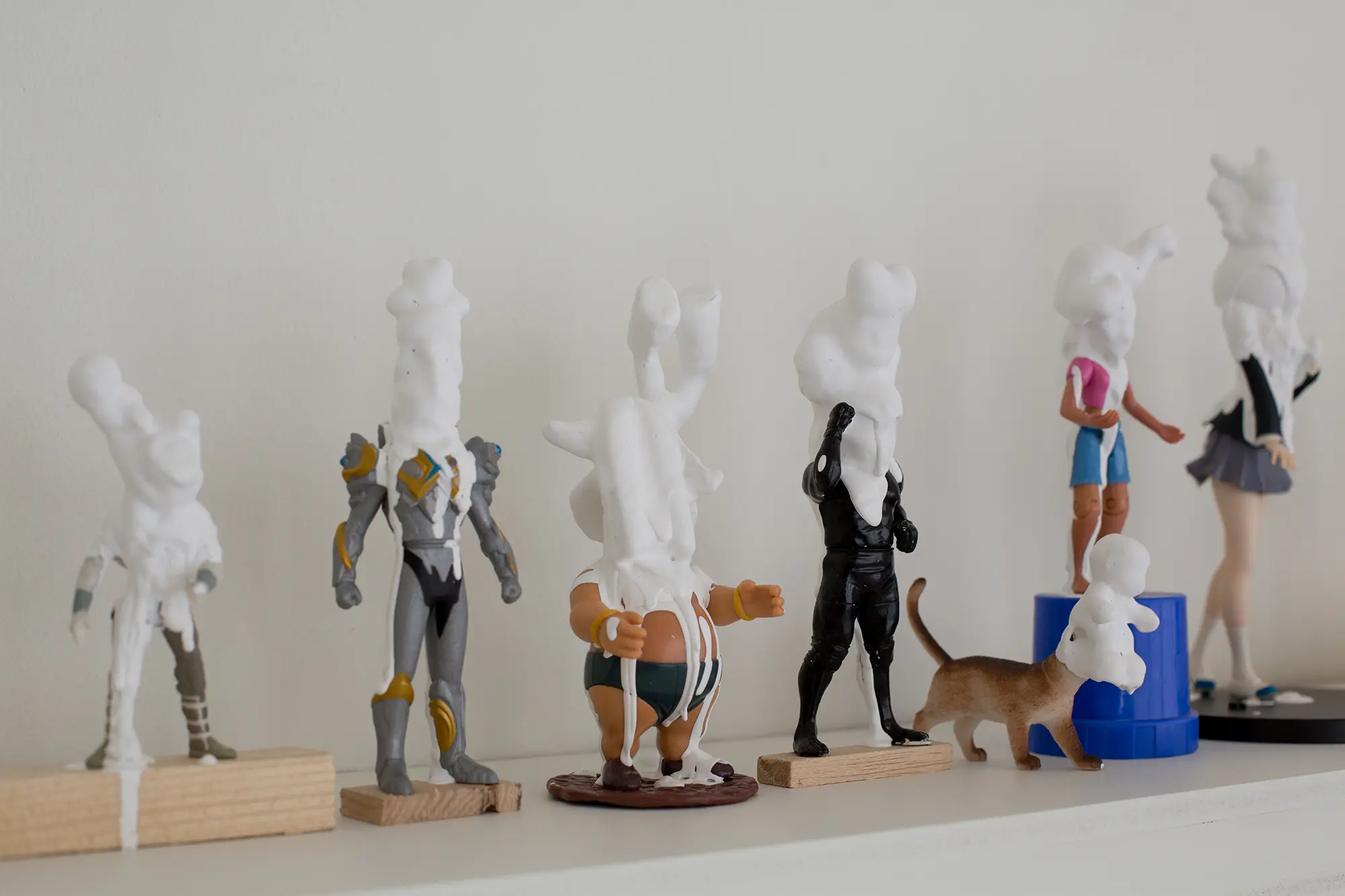 Framför en vit vägg står flera små skulpturer uppställa på rad. Skulpturerna består av leksaksfigurer som delvis är täckta av vit plastmassa som täcker deras huvud.