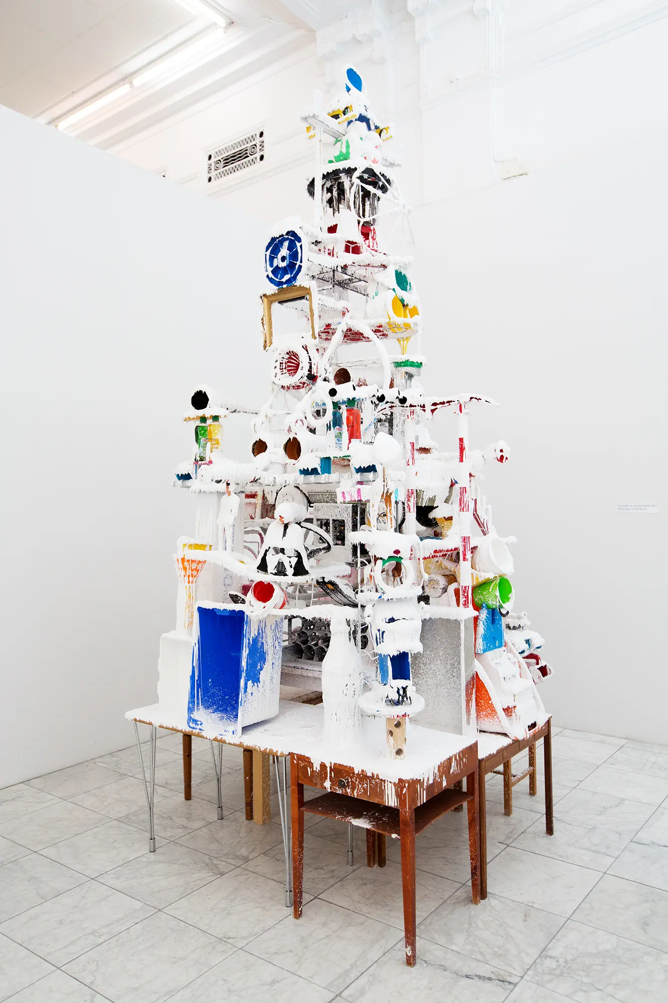 I ett vitt galleri står en skulptur uppbyggd av en mängd olika föremål staplade på varandra i en hög. Föremålen är till stor del täckta av en vit plastmassa. Bland föremålen syns plasthinkar, diskmedelsflaskor och en tavelram. Föremålen står på flera mindre träbord. 