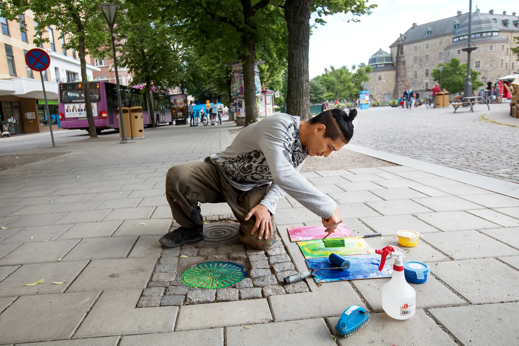På ett torg står en man på knä på stenplattorna och målar ett litet brunnslock i grönt och blått. Bredvid honom står målarverktyg. I bakgrunden syns Örebro slott.