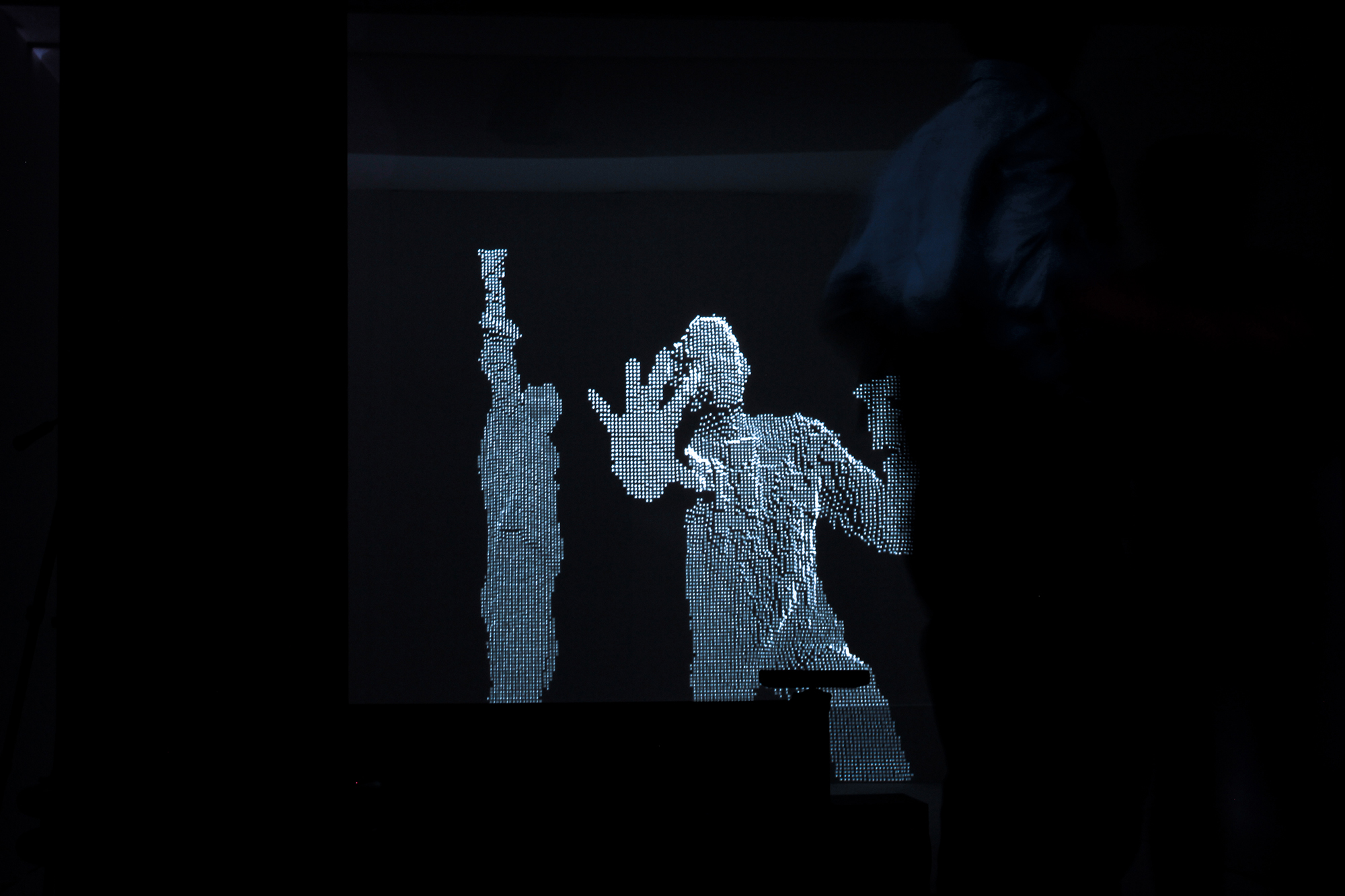 I ett mörkt rum är en mörk skärm placerad som visar två siluetter formade som två människor med blå gröna prickar. Ena siluetten håller fram sin ena hand med en öppen hand vilket signalerar stop. 
