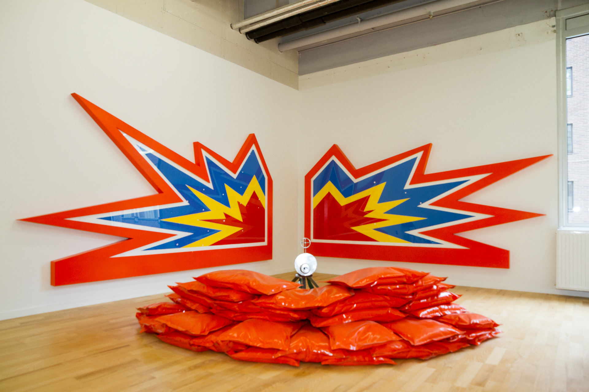 I ett hörn av ett rum står en installation som föreställer en kulspruta i mitten och framför kulsprutan ligger röda säckar på varandra som skapar en liten mur. På vägen i bakgrunden sitter två stora skulpturer som föreställer två explosioner gjorda i serietidnings tecknat format.