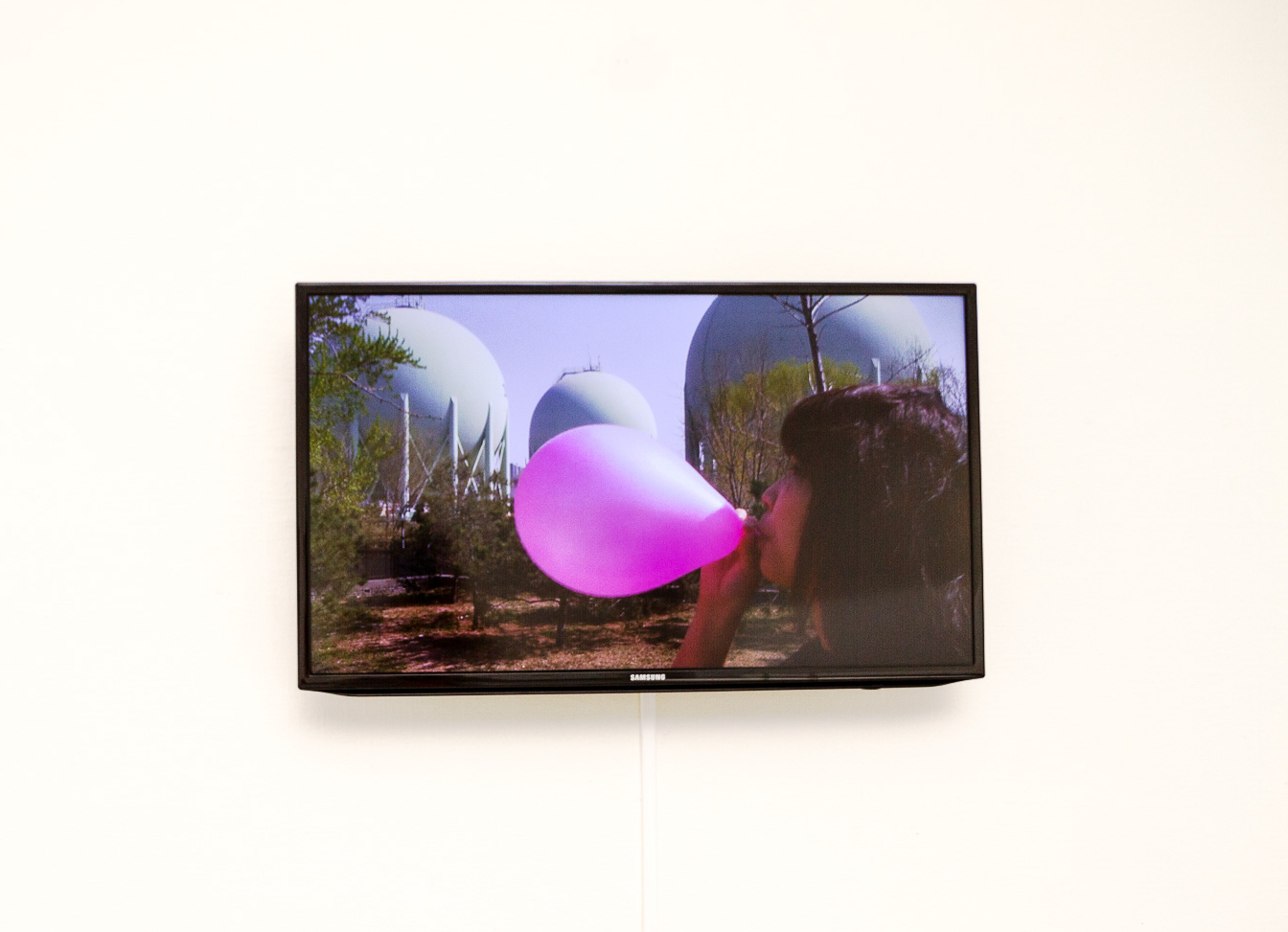 På en vitt vägg hänger en svart TV som visar en bild av en kvinna som står i ett naturområde och blåser upp en rosa ballong. I bakgrunden syns tre stora runda silos som är blåfärgade.