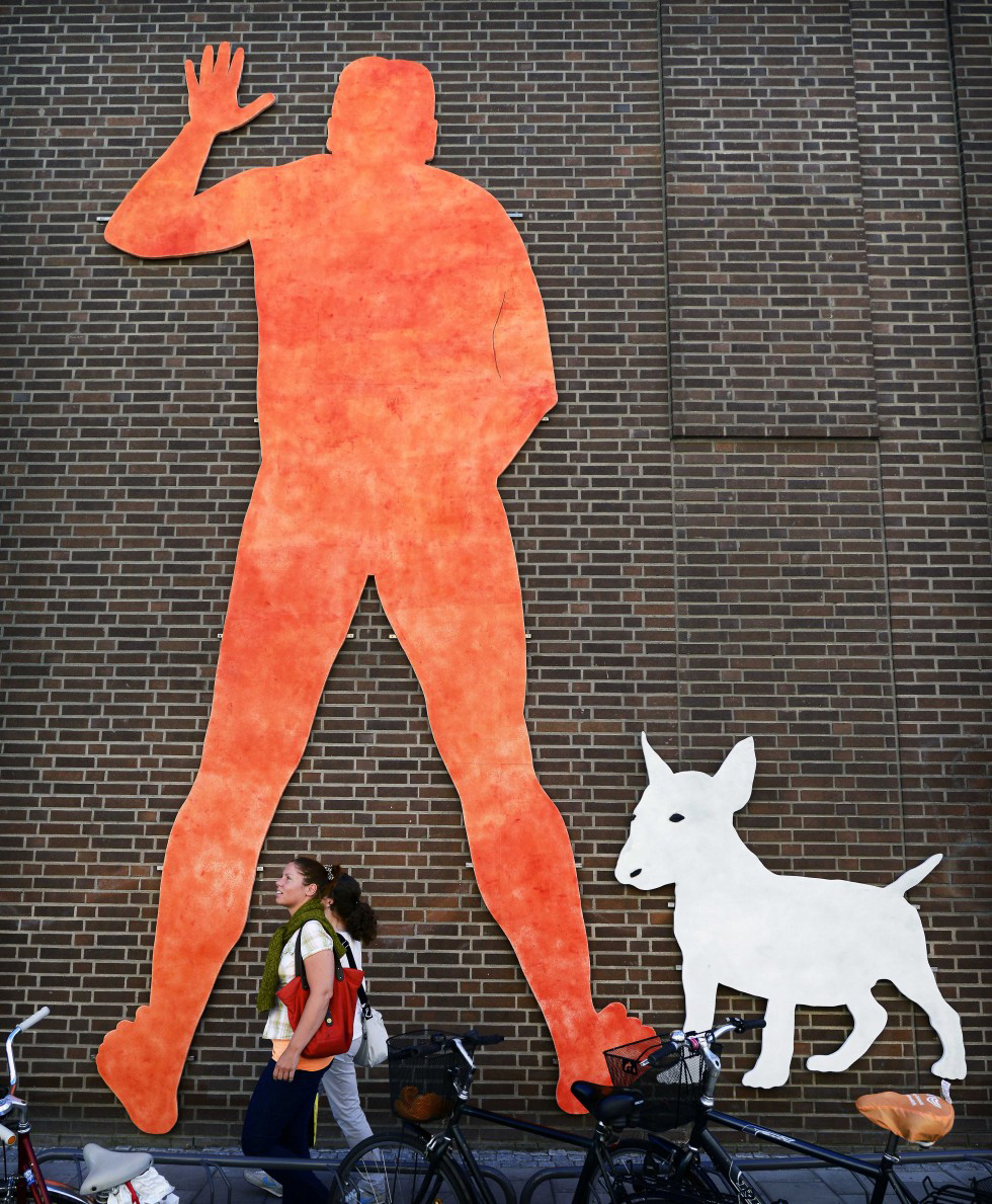 På en tegelfasad hänger en enorm orange siluett av en man som står i en ställning där man kan tro att han kissar. Eftersom man bara ser honom bakifrån med särade ben, ena armen är upptryckt mot väggen och den andra är böjd inåt mott kroppen. Vid mannens ena ben står en vit bullterrier hund.