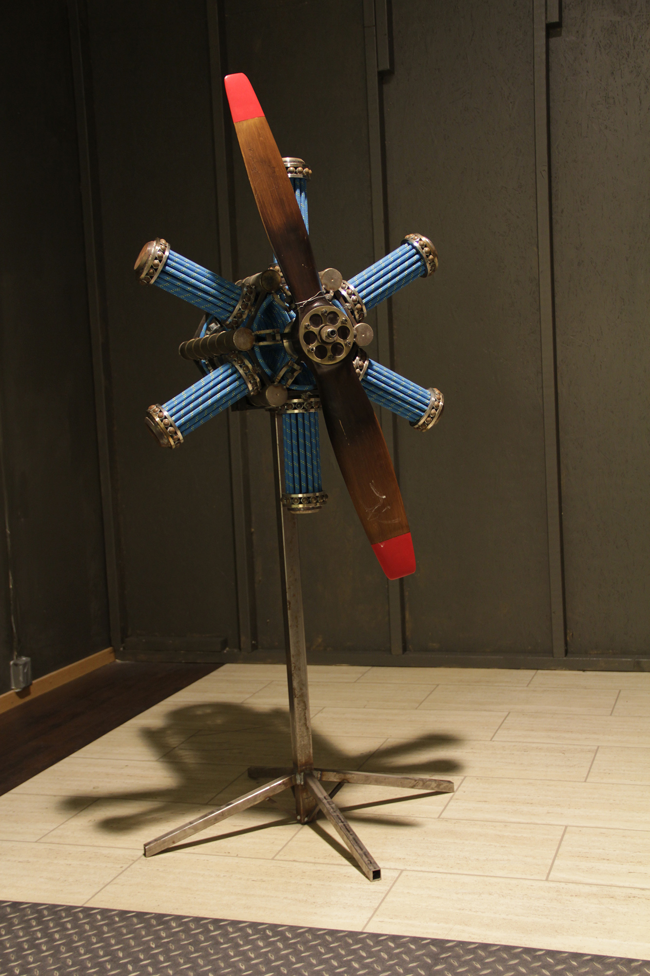 I ett rum på en stålställning sitter en skulptur med sex stycken kapslar i blått rep och i mitten sitter en bronsfärgad propeller med röda kanter. 