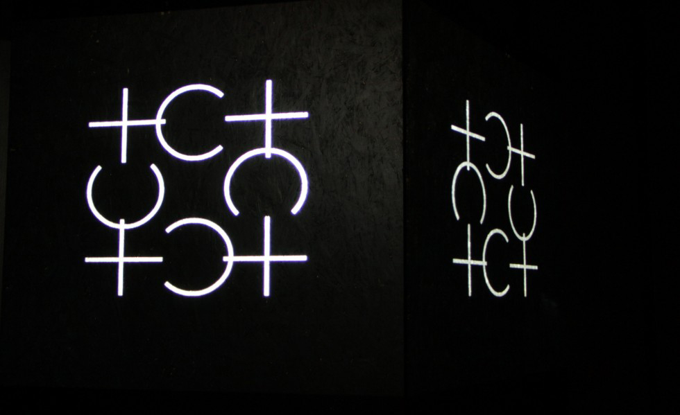 I ett mörkt rum på en svart vägg lyser ett konstverk upp. Konstverket består av många plus tecken och bokstaven C tecken som nuddar varandra. 