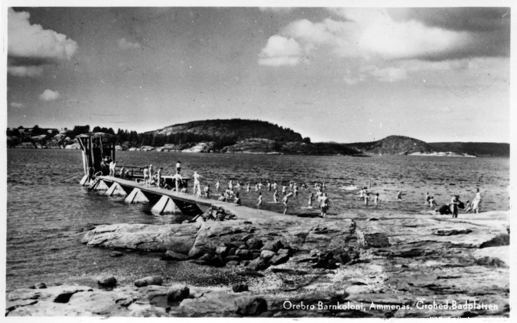 Många barn på brygga vid havet med klippor i bakgrunden. Bad vid Ammenäs barnkoloni i Bohuslän, 1940-tal. Fotograf K. Berg/Örebro stadsarkiv