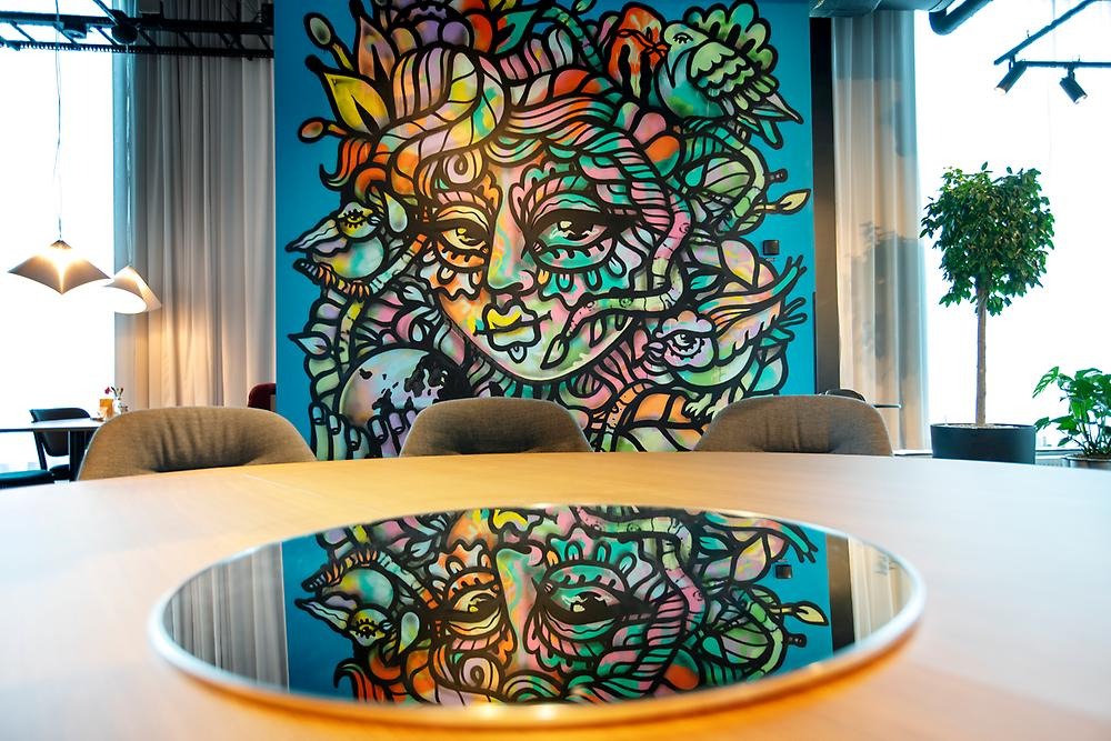 Indoors on a wall in a hotel is a mural of a woman's face painted with colorful colors and black outlines. The hair is decorated with flowers and leaves.