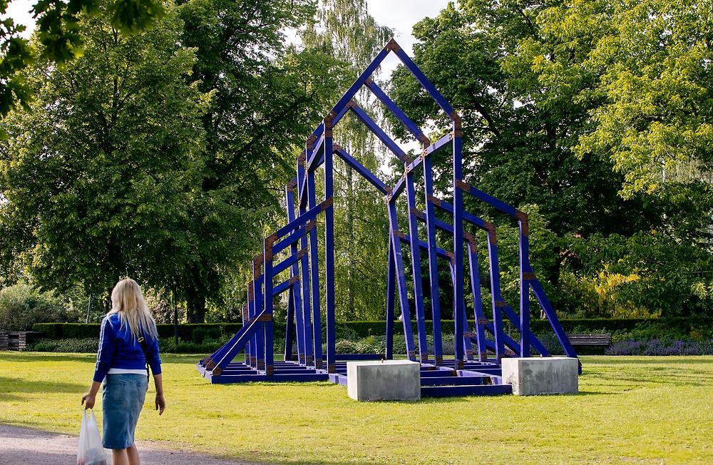 A construction made of blue painted wood, it resembles a church in construction, but there are no walls. It is located in a park environment. A woman walking past the artwork is visible in the image.