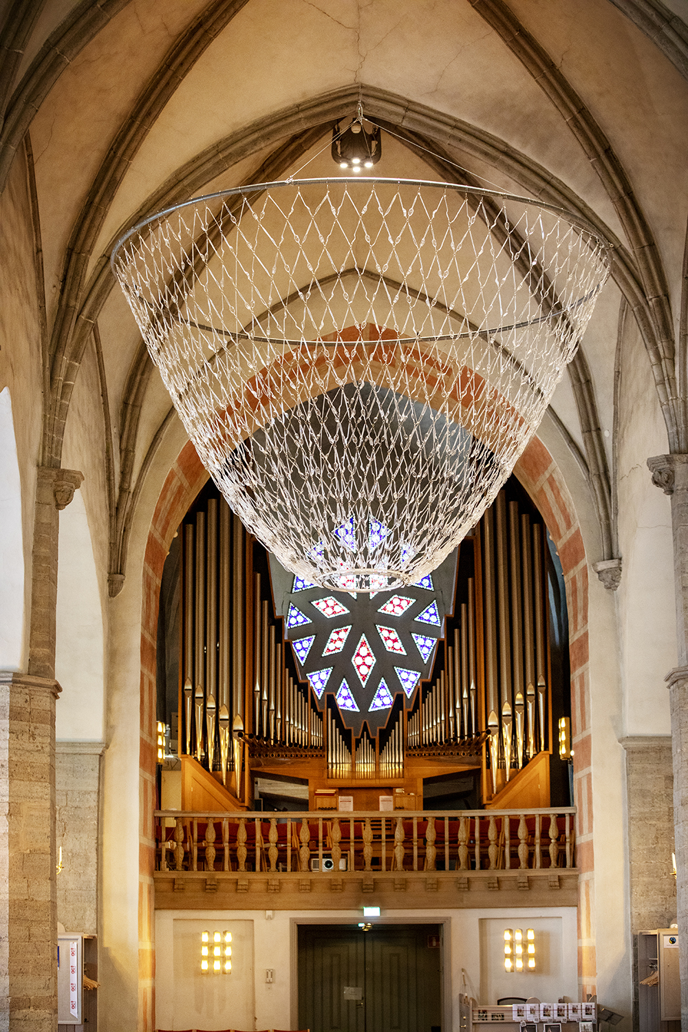 Up in the ceiling inside the Sankt Nicolai church hangs a large chandelier made of transparent cutlery. The crown is shaped like an upside-down round cone and the cutlery is assembled with small threads and rings so that they create a quadrilateral triangle pattern. In the background, an organ can be seen in front of an incredibly colorful window.