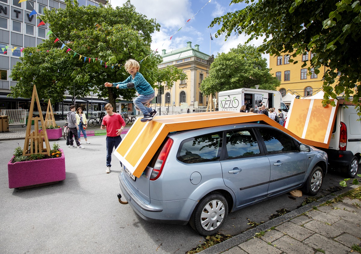 Ett barn hoppar från en springbana som är uppbyggd på bilar som står ute. i bakgrunden syns personer som tittar på. 