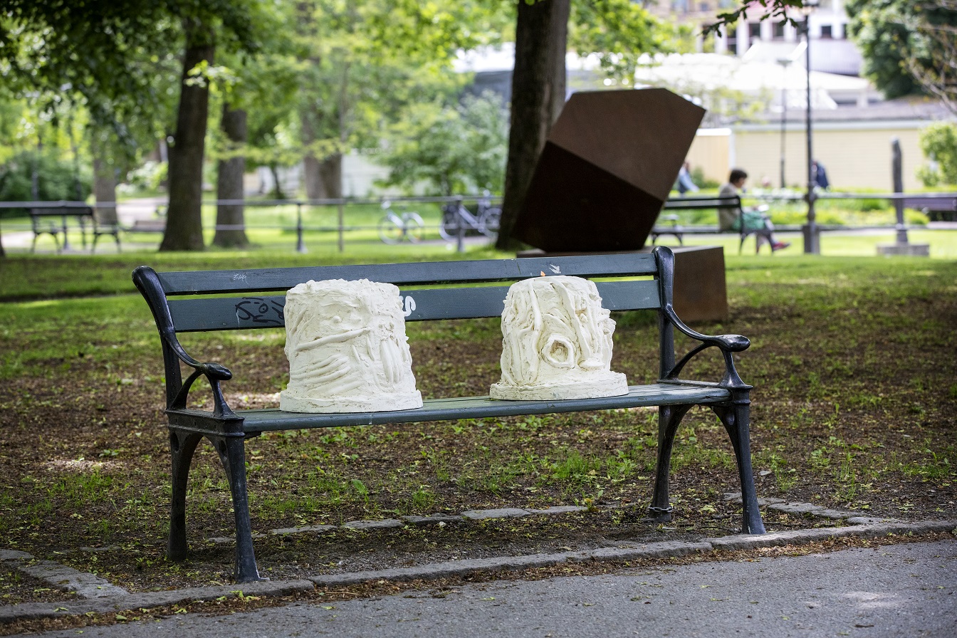 En parkbänk med miljö bakom sig. På bänken sitter två vitgulaktiga skulpturer. I skulpturerna kan man tyda händer som samtalar i teckenspråk.