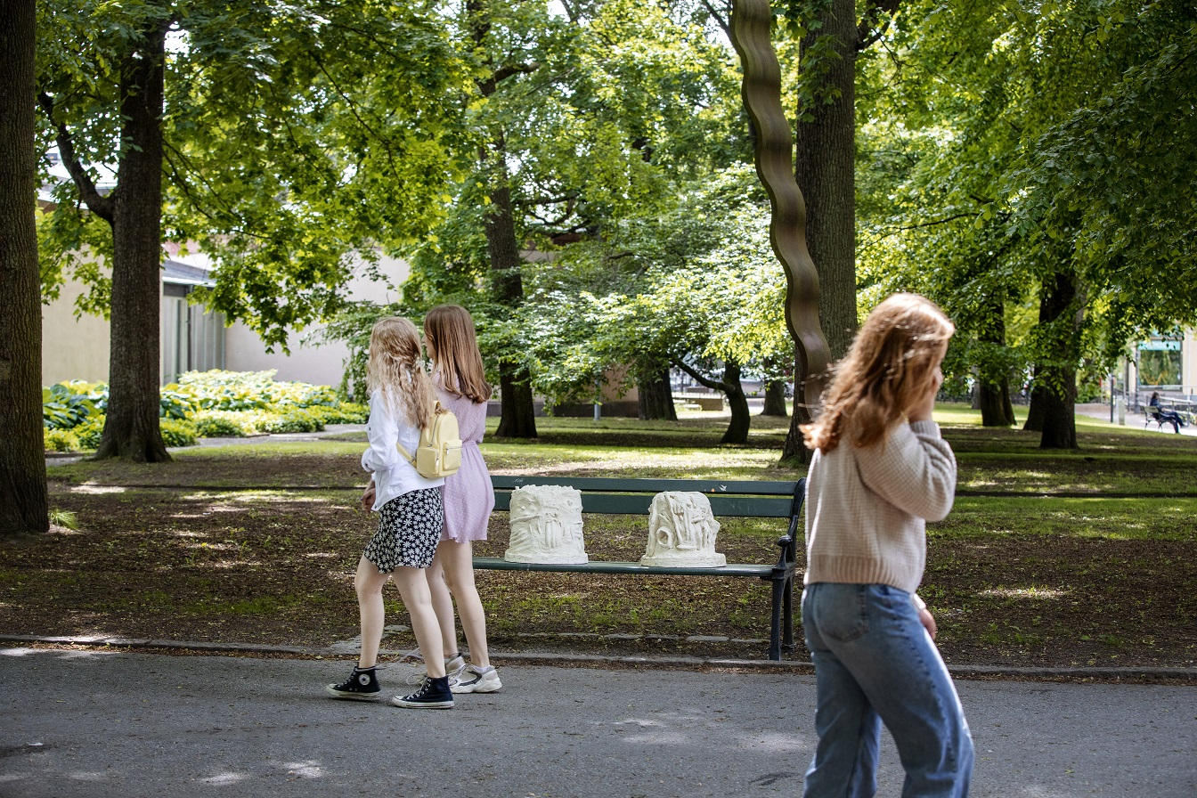 People walk past a park bench with a park environment behind them sit two white and yellow sculptures. In the sculptures, you can make out hands that converse in sign language with each other.