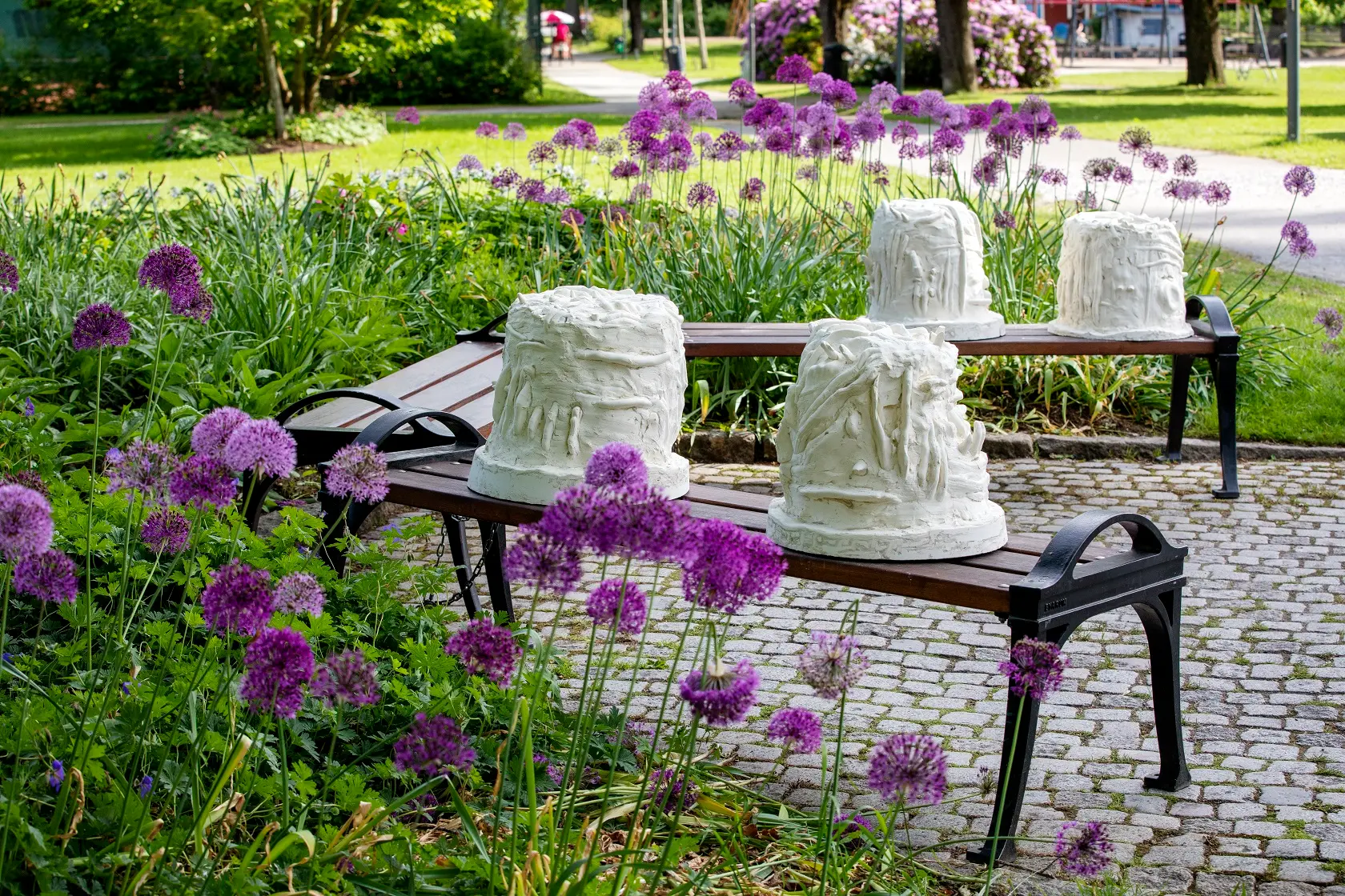 Tre bänkar i stadsparken. På två av bänkarna sitter två vardera vitgulaktiga skulpturer. I skulpturerna kan man tyda händer som samtalar i teckenspråk.