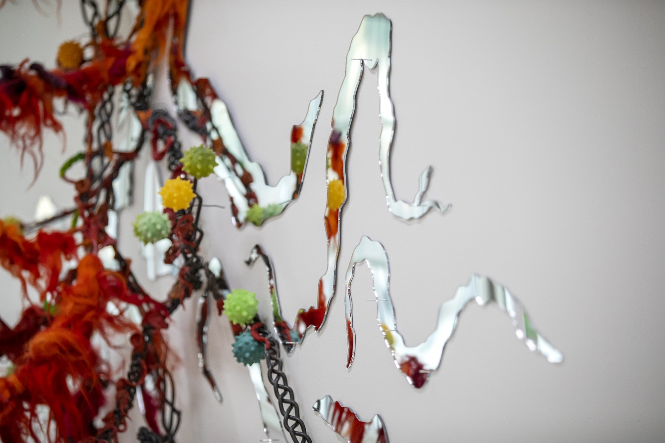 Närbild på skulptur som är färgglad och består av långa grenar av plast, tyg och speglar som klättrar på väggen. 