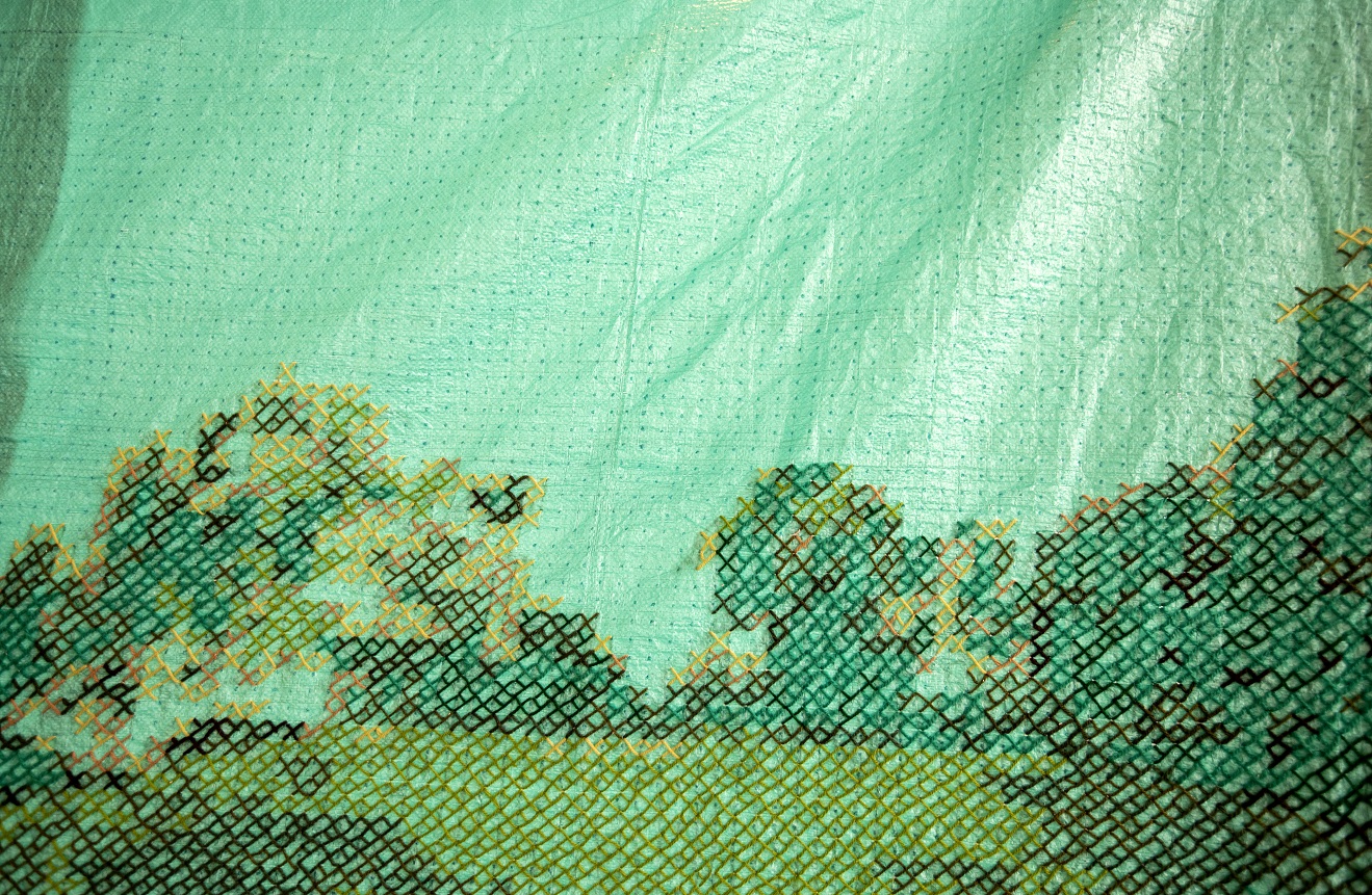 Närbild på konstverket som består av en presenning där korsstygn i  X och Y i gröna nyanser utgör ett landskap. 