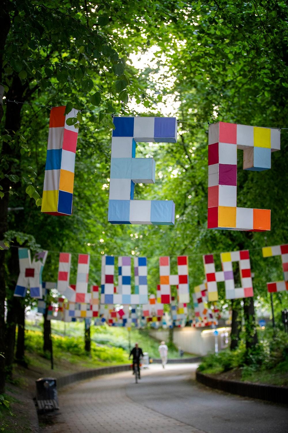 Above a bicycle lane in the trees hang large letters from the Latin, Hebrew and Cyrillic alphabets. The letters are three-dimensional, white and with sewn-on rectangular textile pieces in blue, red and orange tones. In the background is a blurred man on a bicycle.