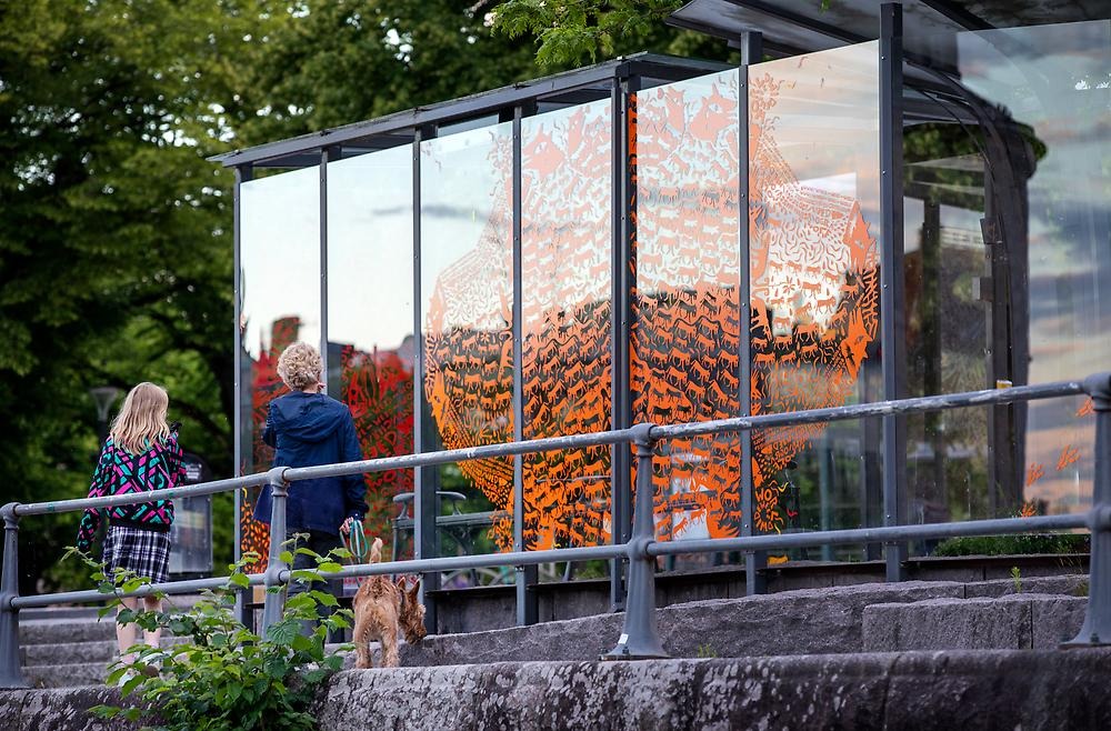 Two people look at a bus shelter whose windows are decorated with orange and red vinyl plastic in the shapes of wolves and various animals. The animals create a pattern on the glass that resembles petroglyphs.