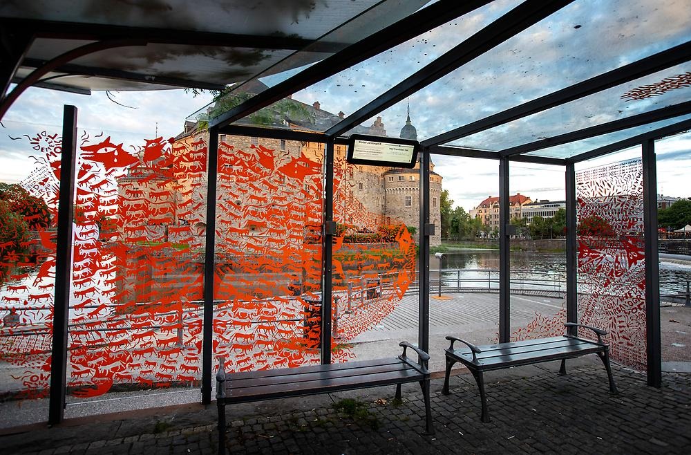 The photo is taken from inside a bus shelter whose windows are decorated with orange and red vinyl plastic in the shapes of wolves and various animals. The animals create a pattern on the glass that resembles petroglyphs. In the background you can see Örebro Castle and the water in front it.