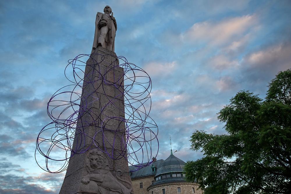 On the Karl XIV Johan statue hangs a cluster of purple rings, the picture is taken from below and in the background you can see a dramatic sky and a tree.