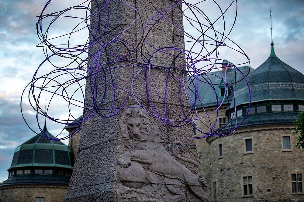 Close-up of the statue of Karl XIV Johan, which has been decorated with purple rings hanging around the statue. On the statue's foundation you can also see a lion which is part of the statue and in the background you can see part of the castle.