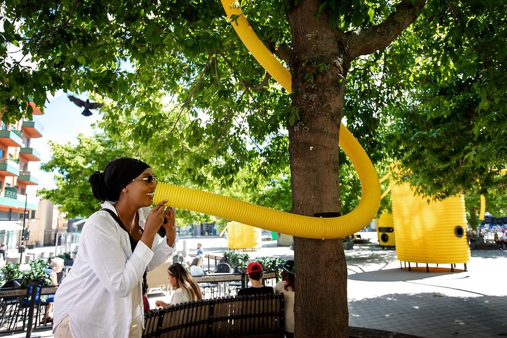 A woman wearing sunglasses, a veil and a white shirt looks happy as she talks into a yellow drainage pipe in a square setting. The pipe comes from above in the picture and is wrapped around a tree. In the background you can see the square with the artwork in the background.