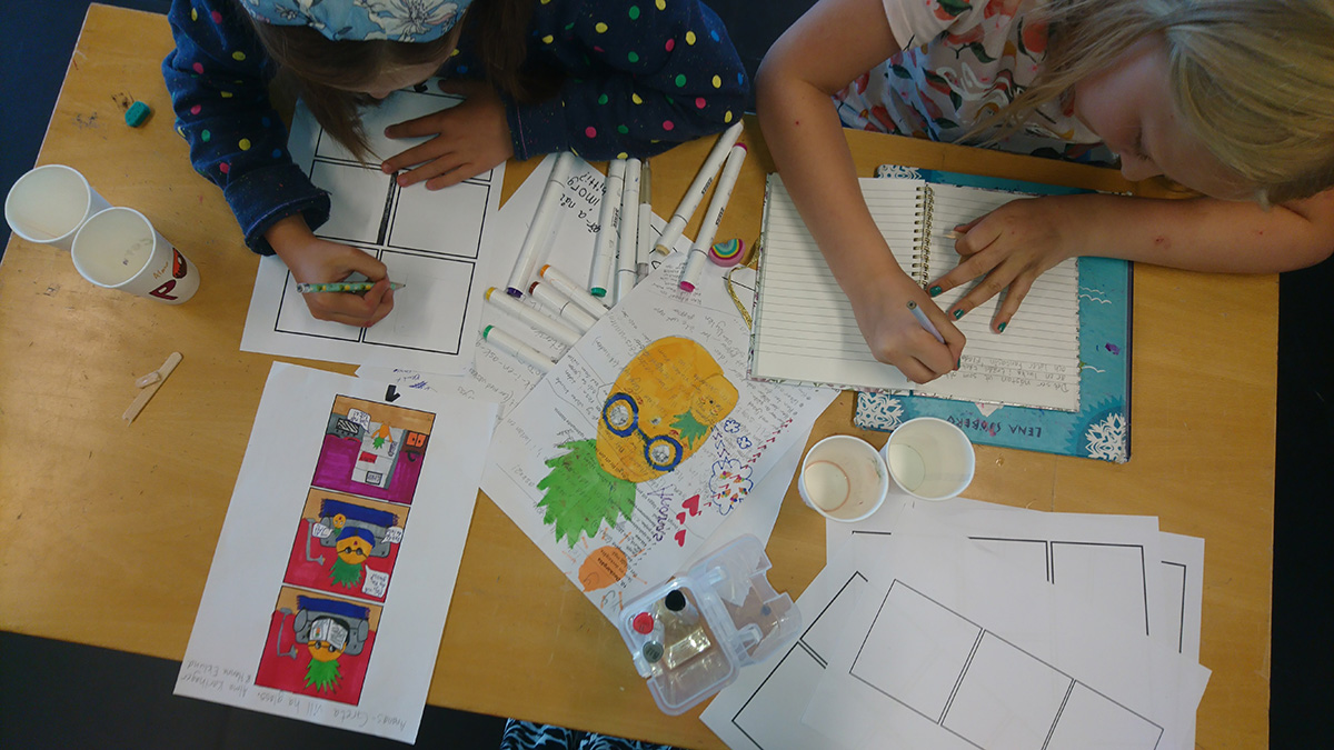 Bilden är tagen ovanifrån ner på två barns som skriver i ett kreativt kaos. Papper med textuppgifter är utspridda på bordet.