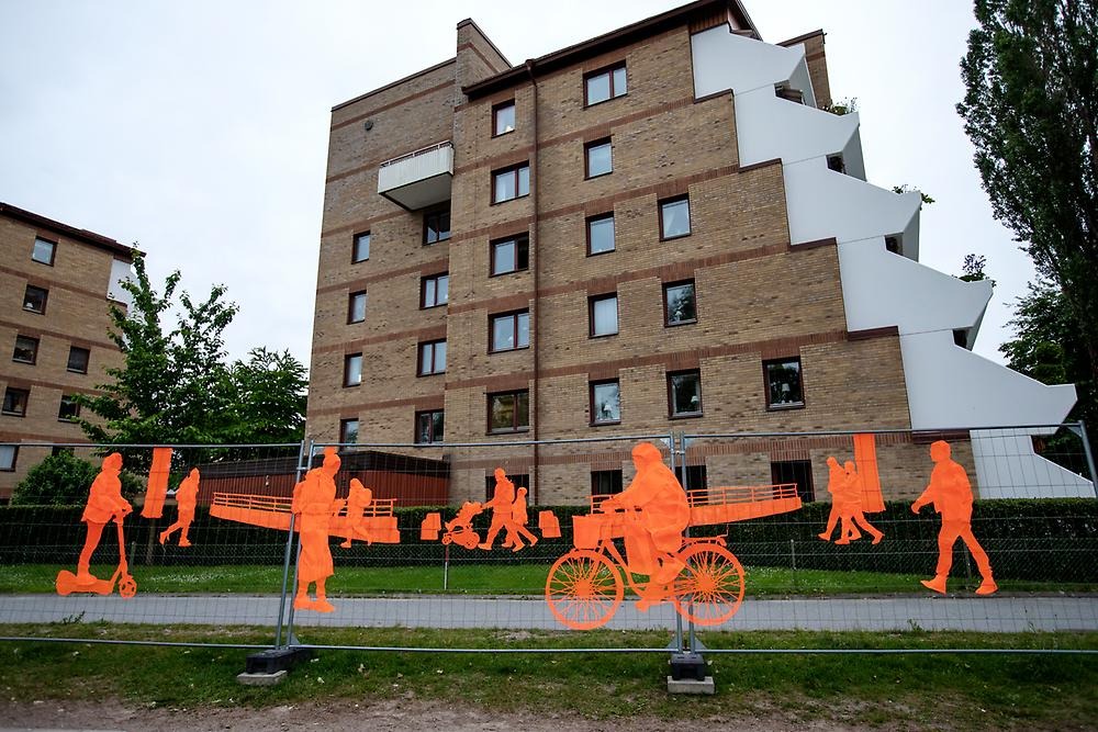 Ett högt staket följer promenadstråket vid Svartån mot stadsparken. Staketet består av orangea broderier som avbildar människor i farten. Människorna cyklar, går samt åker el sparkcykel. 
