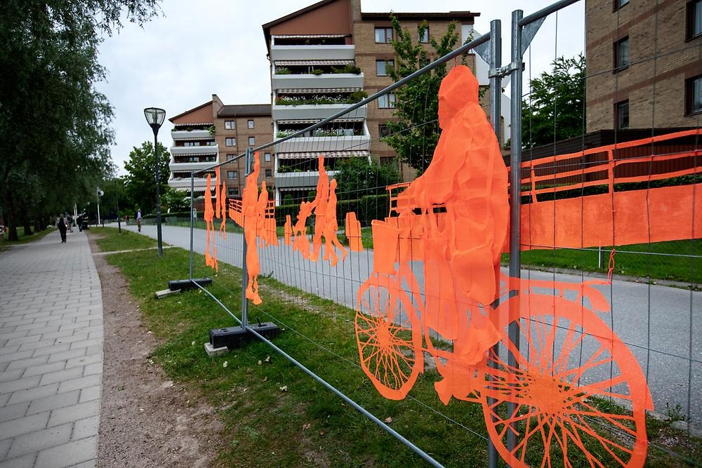 Ett högt staket följer promenadstråket vid Svartån mot stadsparken. Staketet består av orangea broderier som avbildar människor i farten. Människorna cyklar, går samt åker el sparkcykel.