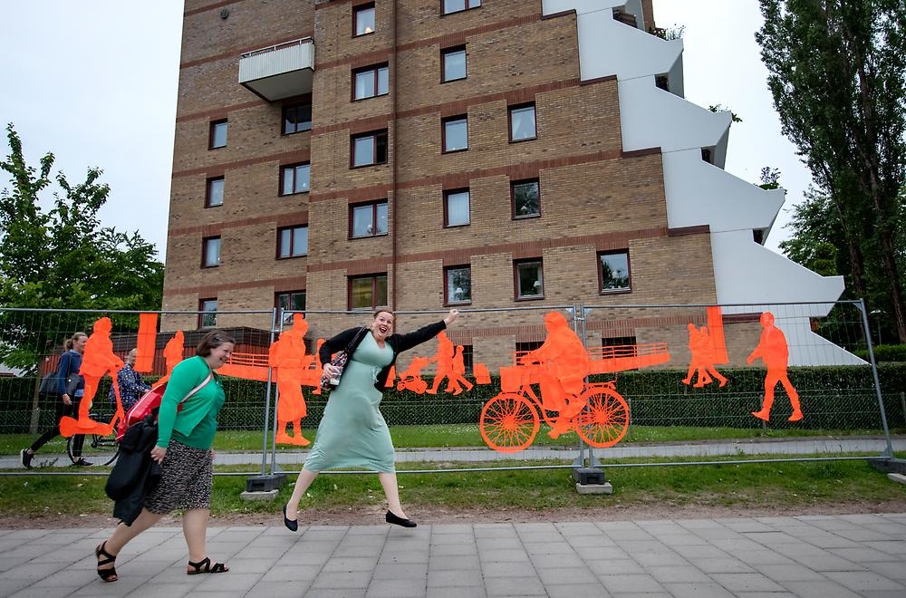 Två kvinnor skrattar och ena hoppar framför det stora staketet som följer promenadstråket vid Svartån mot stadsparken. Staketet består av orangea broderier som avbildar människor i farten. Människorna cyklar, går samt åker el sparkcykel. 