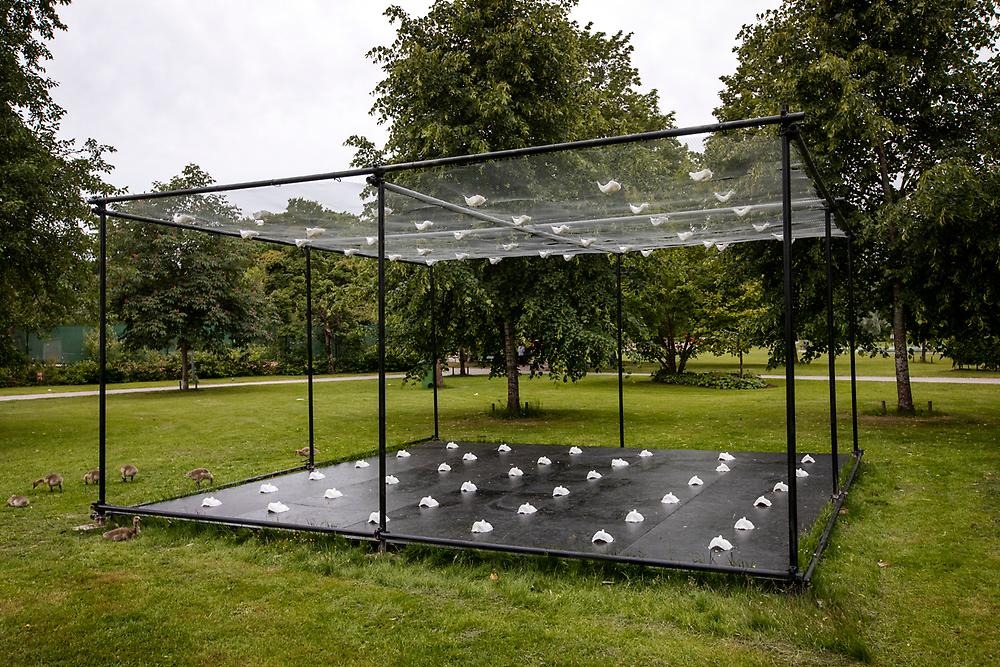 On a patch of grass in the city park stands a black stand with a black base and a white net on top. On the foundation are white face masks that look up and meet white face masks hanging in the net that looks down.