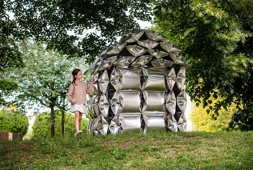 In a leafy park stands a house made of high-gloss aluminum pillows. The surroundings are reflected in the cushions and a girl stands next to the house/sculpture.