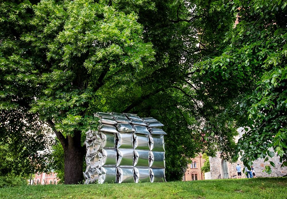 In a leafy park stands a house made of high-gloss aluminum pillows. The surroundings are reflected in the cushions.