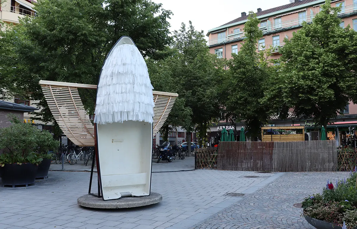 Outside at Järntorget, an old boat can be seen standing upright. On the sides of the boat, wooden wings stick out and on the top, white rustling fabric hangs down over half the boat in a material similar to pom pom balls fixed oblong.