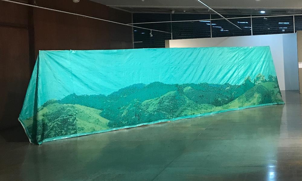 In an empty room, a large green tarpaulin can be seen hanging over a high fence. The tarpaulin is decorated with cross-stitches in shades of green that form a landscape with mountain-like shapes.