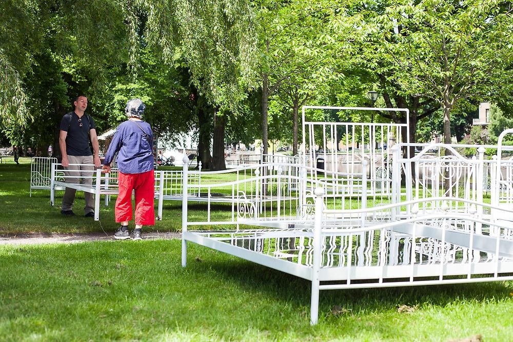 White iron bed frames stand on a green lawn and where the mattress is usually placed is instead a mirror. Two people carry a bed to position it correctly.