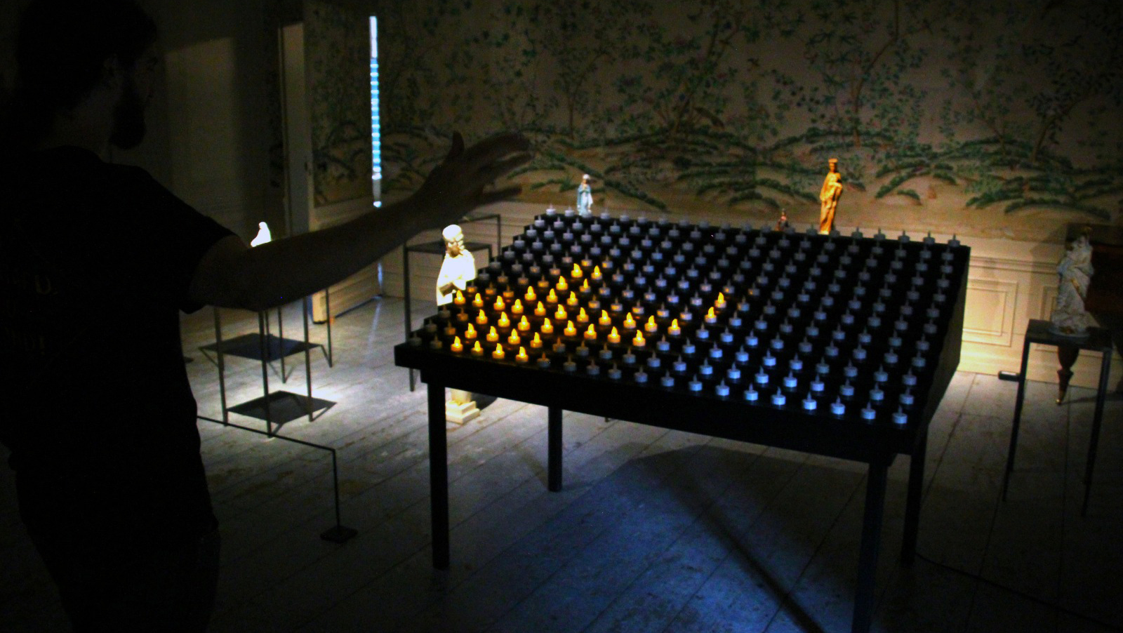 A man stands with outstretched hand in front of an altar table filled with prayer candles, the candles are ordinary battery candles and only a few of them in a corner shine in the pattern of a cross. In the background are black pedestals with various church statues on them.