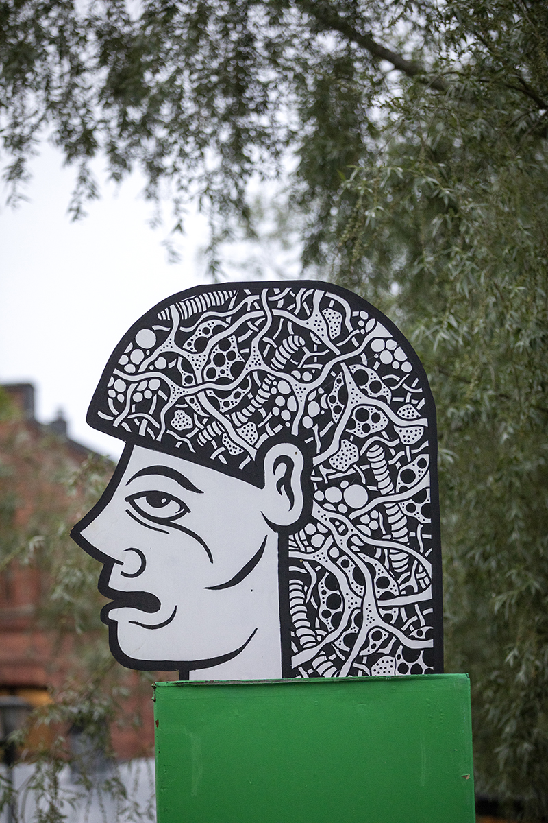 A close-up of an illustrative face in black and white where the person's hair is painted in various graphic patterns is placed atop a green painted pillar.