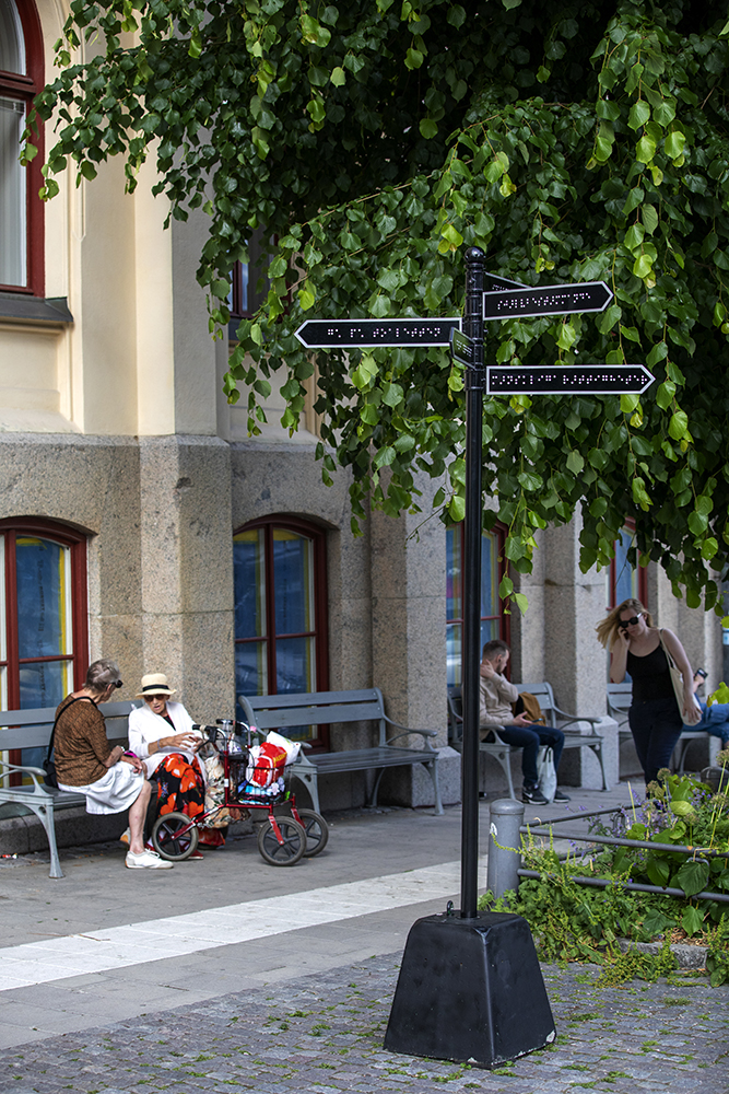 In the foreground of the City Hall in Örebro, a sign is used that is usually used as a directions sign. Instead of the Latin alphabet, Braille can be seen on the signs. The sign is black with an arrow formation in the front edge. A white border can be seen around the outer edges of the sign. The sign's pole and foundation are black. The picture shows the sign in its entirety, behind the sign you can see some people sitting down on a park bench in front of City Hall. On the right edge you can also see the shrubbery from a tree with green leaves.