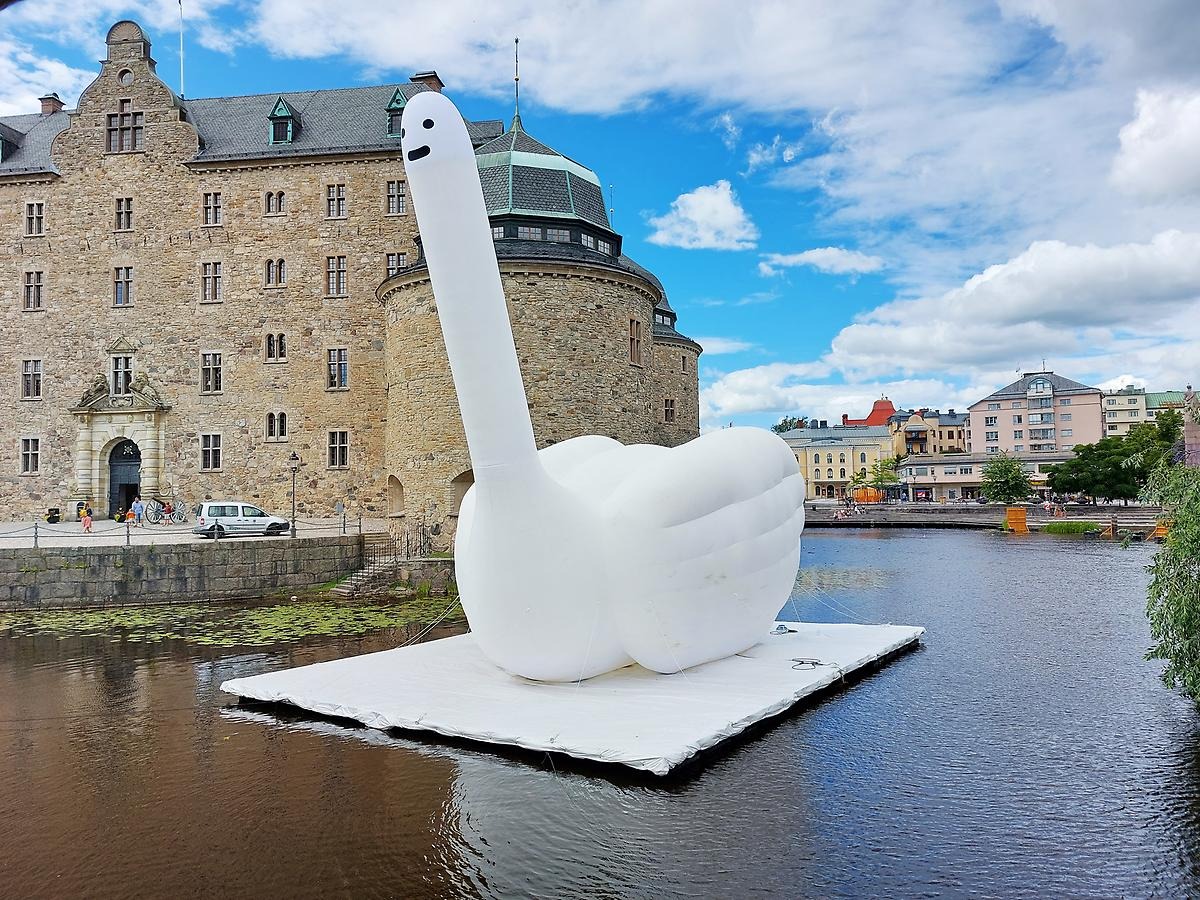 A 12 meter high inflatable white swan floats on the water in front of the castle's grand entrance. The swan has no beak and an untroubled blank expression, two black dots as eyes and a black line as a mouth. The swan's neck is shaped like an middle finger and goes straight up towards the sky, and the body has two folded wings on either side.